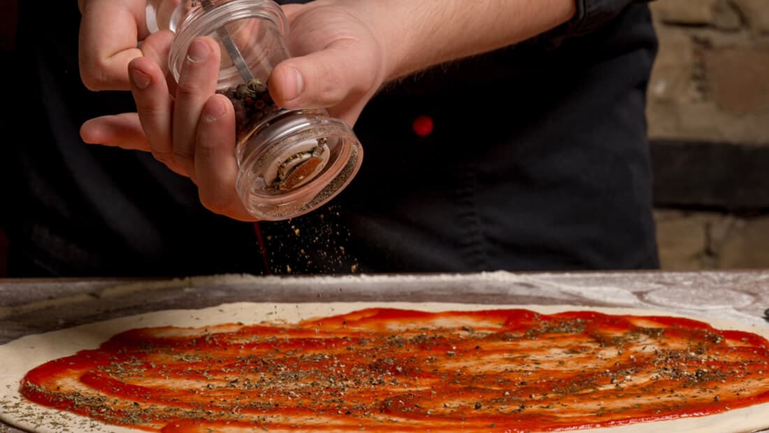 A man is spreading pepper on a pizza covered with tomato sauce
